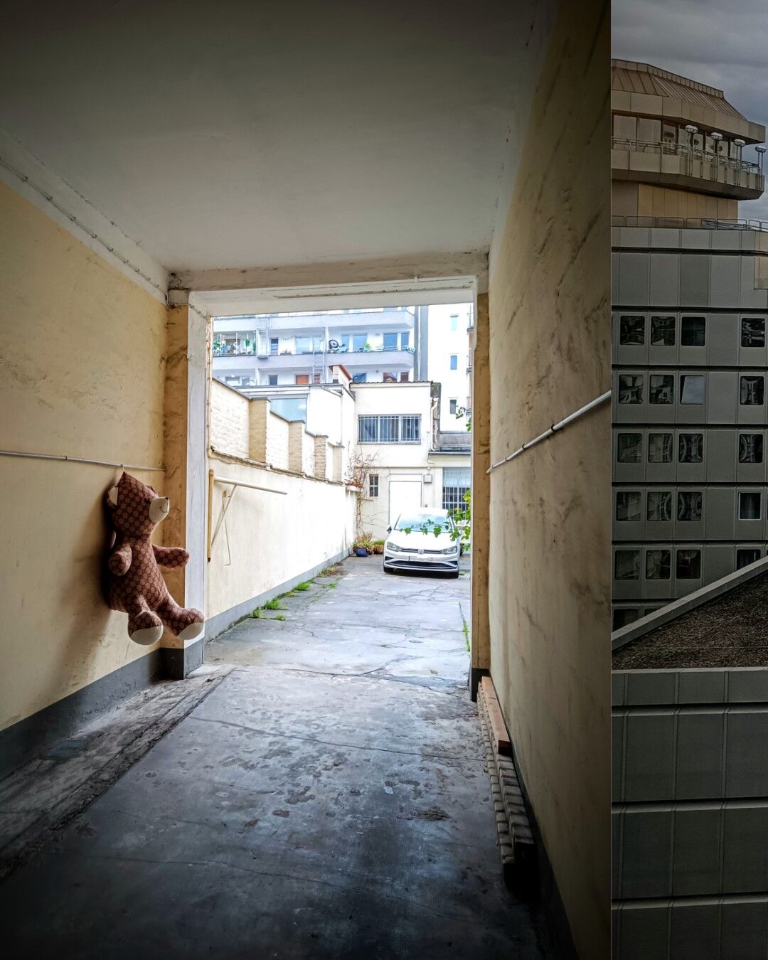 The image shows a narrow passageway with beige walls and a concrete floor. On the left wall, there is a large teddy bear hanging by its arms. The passageway leads to an open courtyard where a white car is parked. In the background, there are several buildings with balconies and windows. The right side of the image shows a partial view of a modern building with a grid of windows and a rooftop structure.