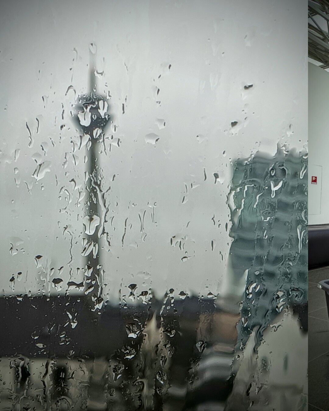 A blurry view of a cityscape seen through a rain-drenched window. Water droplets distort the scene, which includes the Düsseldorf Rhine Tower (broadcast tower) in the distance, partially obscured by the rain. In the foreground, the windowpane is covered with numerous small drops of water. The sky appears overcast and gray, with a faint hint of buildings in the background. The image has a soft, diffused light and an overall melancholic, rainy atmosphere.