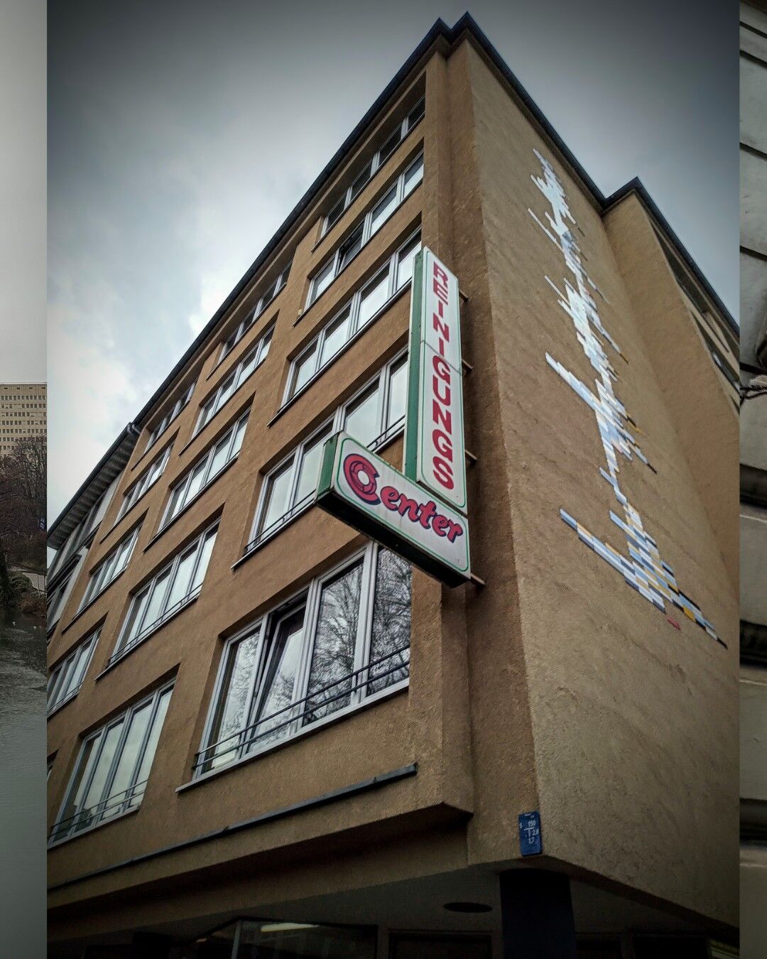 The image shows a close-up view of a building's corner, taken from a low angle. The facade is a beige-colored wall with rows of windows. A neon sign, reading "REINIGUNGS CENTER," is attached to the building, its bright red and green colors contrasting with the muted tones of the structure. To the right of the sign, a mural-like design stretches vertically along the building's side, with abstract shapes and lines in white and other colors, resembling a broken, pixelated pattern. The sky above is cloudy, adding a gray atmosphere to the scene.