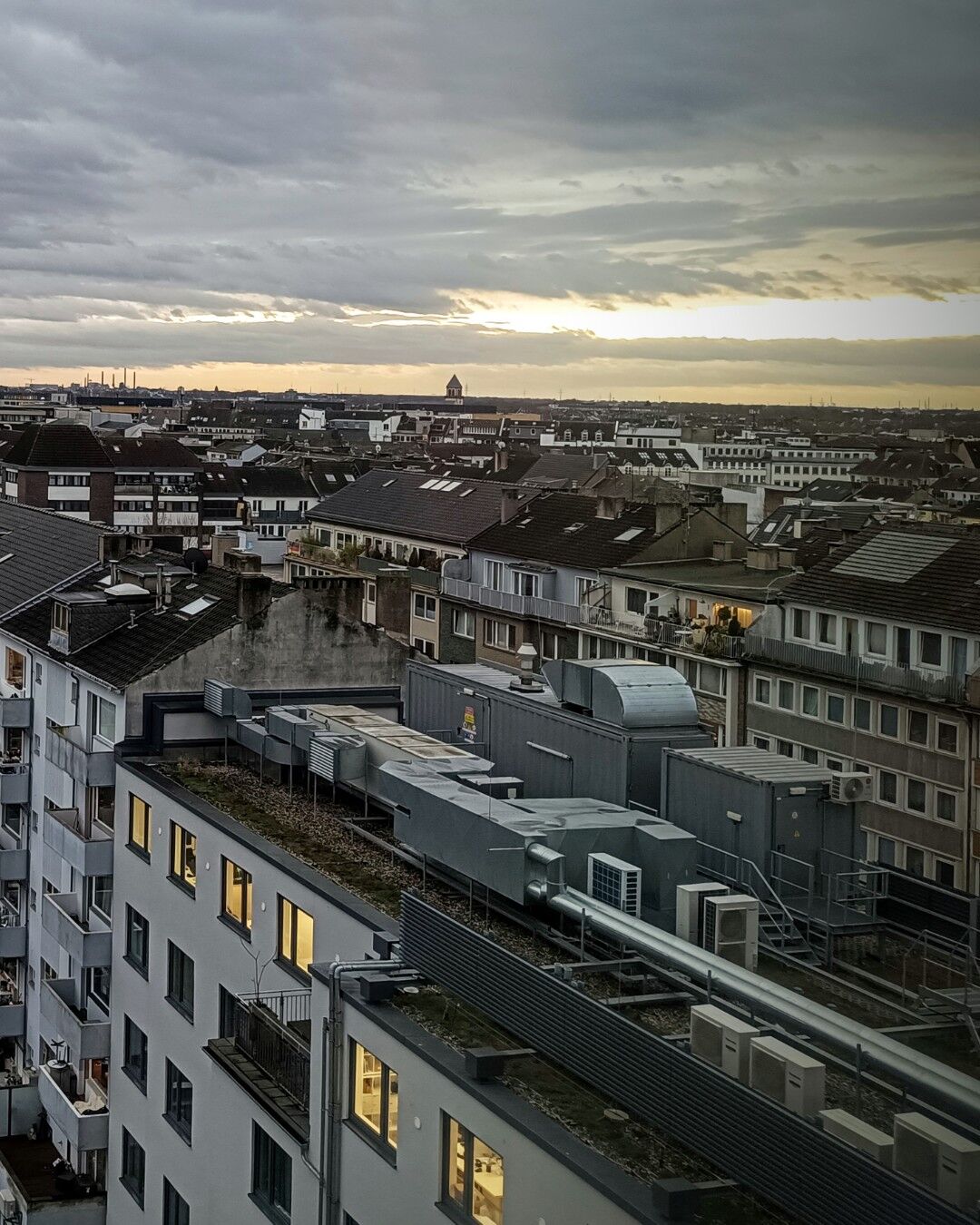 (Image part 2 of2) The image shows the urban cityscape of Düsseldorf from Oststraße towards main station with multiple buildings and rooftops. The buildings are primarily modern with flat roofs, some of which have visible HVAC systems and other rooftop equipment. The sky is overcast with thick clouds, but a hint of morining sunlight is breaking through on the horizon, creating a contrast between the dark clouds and the lighter sky. The scene is taken from one of the top floors of the Moxy hotel, offering a wide view of the city. The image captures the density and architectural variety of the urban environment, making it interesting for its depiction of city life and infrastructure.