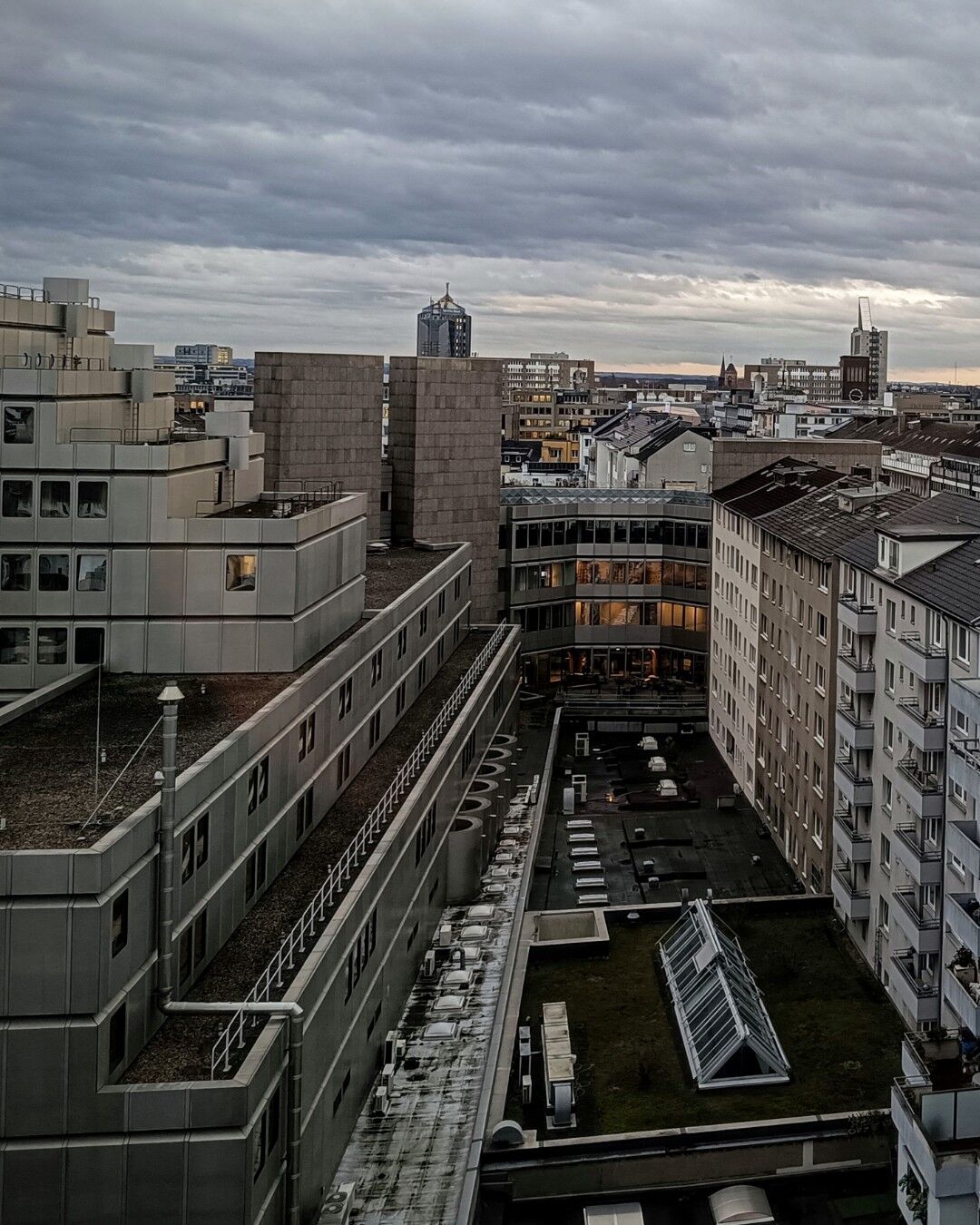 (Image part 1 of2) The image shows the urban cityscape of Düsseldorf from Oststraße towards main station with multiple buildings and rooftops. The buildings are primarily modern with flat roofs, some of which have visible HVAC systems and other rooftop equipment. The sky is overcast with thick clouds, but a hint of morining sunlight is breaking through on the horizon, creating a contrast between the dark clouds and the lighter sky. The scene is taken from one of the top floors of the Moxy hotel, offering a wide view of the city. The image captures the density and architectural variety of the urban environment, making it interesting for its depiction of city life and infrastructure.