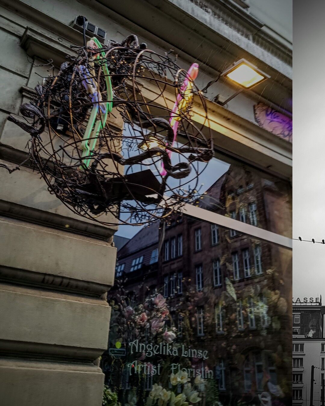 The image shows a decorative, sculptural installation mounted on the exterior of a building. The structure is a large, spherical frame made of twisted vines and metal, with vibrant neon lights in pink and green forming abstract shapes within it. A bright light fixture is attached to the wall above the installation. Reflected in the window below the artwork is a glimpse of a brick building with rows of windows and floral decorations in the foreground. Through the window, a sign is visible that reads "Angelika Linse, Artist Floristin" in green text. The overall scene has a warm, artistic atmosphere, with a mix of urban and nature-inspired elements.