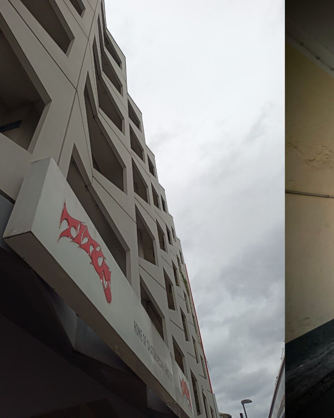 The image shows a tall building with a unique architectural design featuring multiple rectangular windows and balconies. The building has a sign with red text and graphics, with the words 'TITUS'. The sky is overcast with grey clouds, and there is a streetlamp visible at the bottom of the image. On the right side of the image, there is a partial view of the next image an interior wall with some discoloration and a pipe running along it.