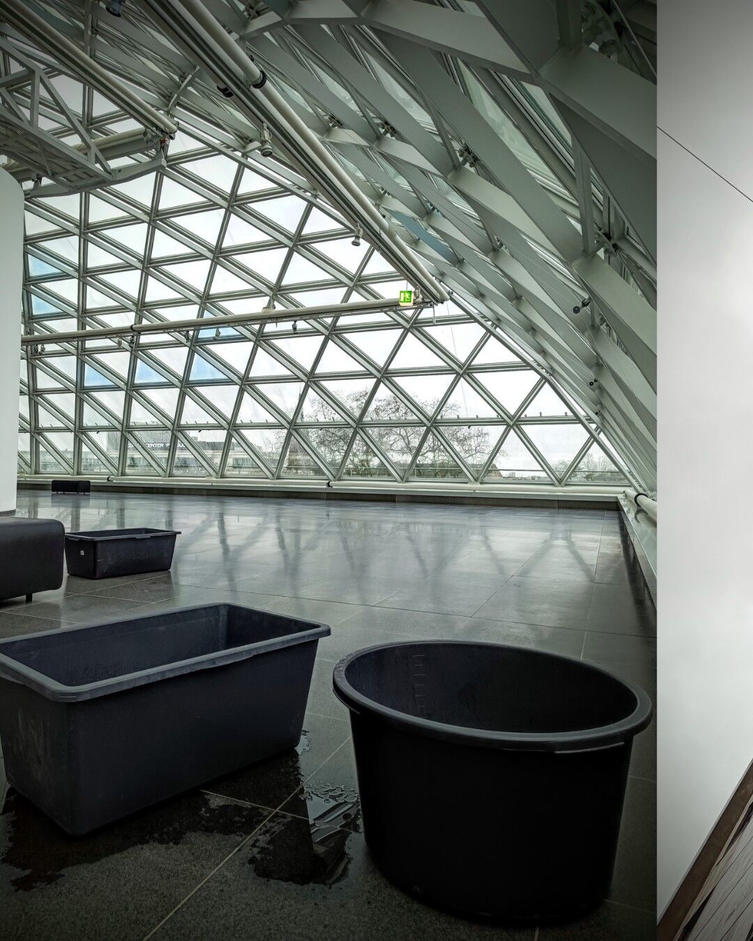 Thr empty, spacious interior of the K21 art museum under the roof with a modern, glass-paneled ceiling featuring triangular shapes. Three black plastic containers, one rectangular and one oval, sit on the shiny gray floor, with water pooled around them, catching leaks from the roof. The background shows a cloudy, overcast sky through the glass, with some trees visible outside. The space feels quiet and minimalistic, with a sense of industrial design highlighted by the exposed beams and clean lines of the architecture.