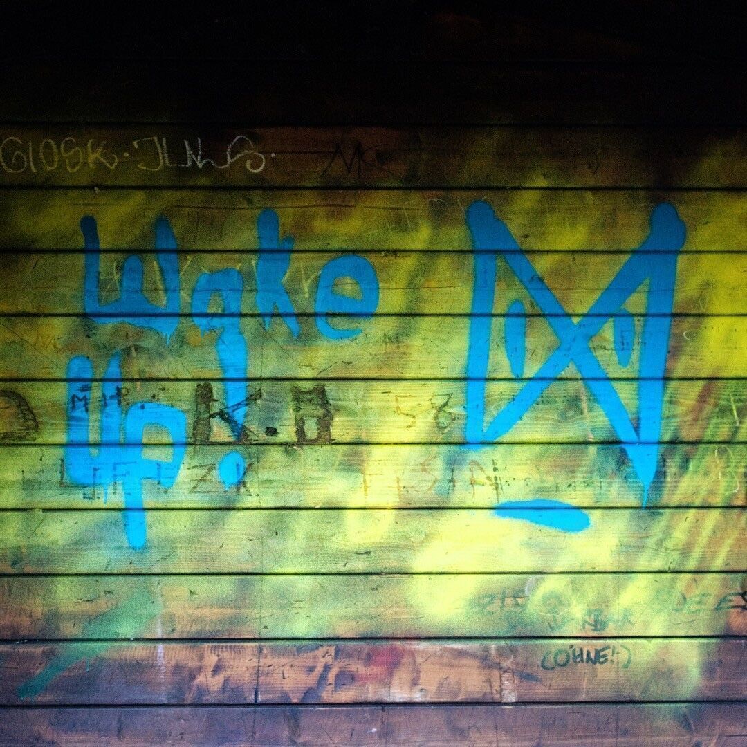 Ein Holzunterstand, der gelb bemalt ist. Darauf eine seitlich gedrehte aufgemalte Eieruhr in blauer Farbe. Daneben in blauer Schrift der Text "Wake Up".