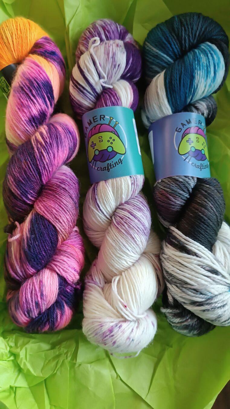 Three hanks of hand-dyed yarn on a light green paper. The hanks are variegated. The left one shows purple, pink and orange. The middle one is mainly natural white with purple parts. The right one is white, blue and anthracite. The middle and right Hank are wrapped with a banderole showing a gamepad and a skein of wool in the background - the logo of the dyer Gamercrafting.