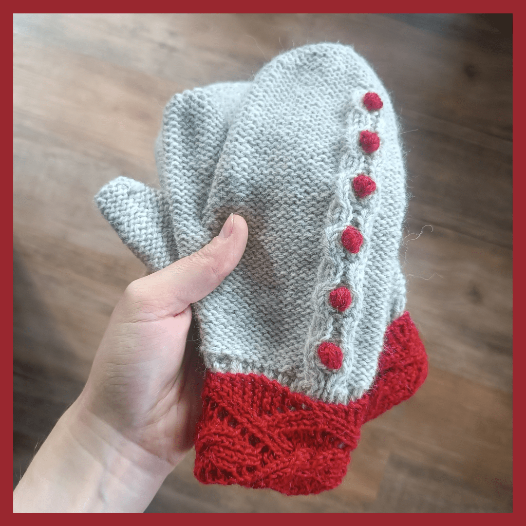 The picture shows a pair of hand-knitted woolly mittens in light grey with red accents. The base is purled with a 1x1 ribbing. The cuff is folded over the ribbing and shows a lace pattern in red. There's also a stripe with waves and red bobbles from cuff to top. The background of the picture is a dark wooden floor and there's a red border around the picture.