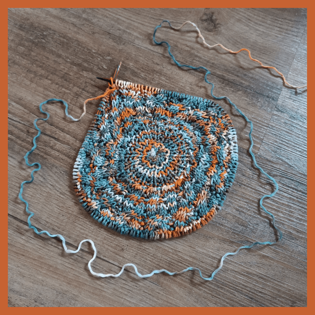 The picture is surrounded by a terracotta coloured border. It shows a blanket, started from the center and knitted in the round. It's about 20 cm across at this stage. The pattern shows cables radiating from the center but the yarn colours make it a bit hard to see them. The yarn is variegated in terracotta, greyish blue and white. The working thread is placed in a circle around the blanket and vanishes in the upper right corner.