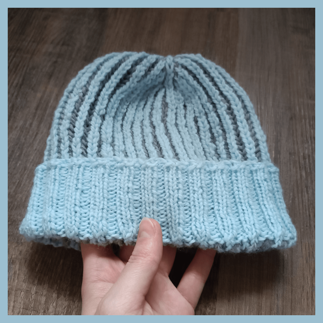 A light blue border surrounds a picture of a light blue and dark grey knitted hat. It's made in two coloured brioche. The main colour is light blue and the turned up 2x2 ribbing is light blue as well. The hat is held by a hand from the down side of the picture in front of a dark wooden floor.