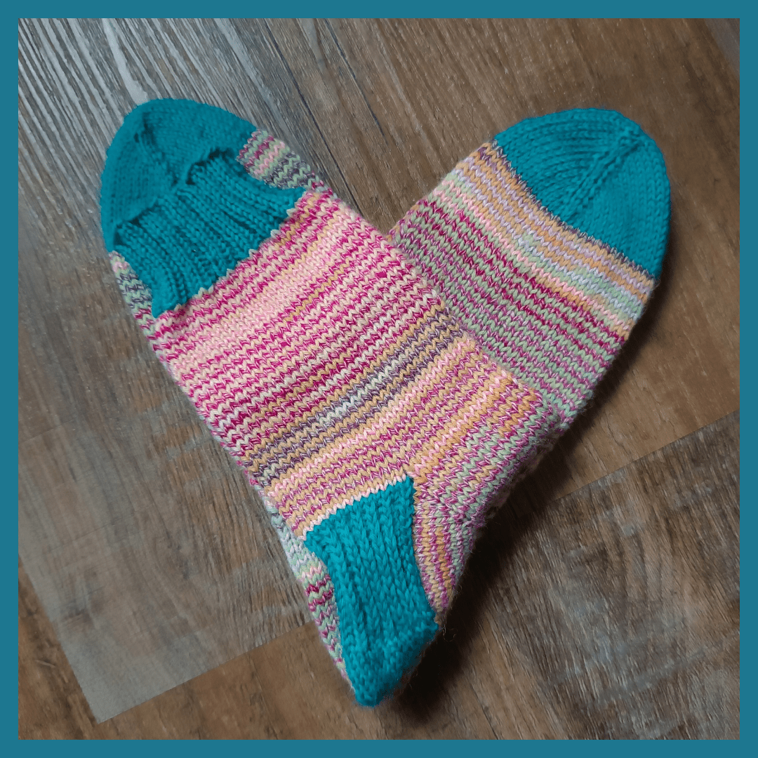A pair of hand-knitted woolly socks forms a heart in the middle of the picture. They lie on a dark wooden floor and there's a teal coloured frame around the picture. The socks have toes, heel caps and a 2x2 ribbing in the same teal colour. The rest of the socks is striped with two variegated yarns. One of them has pink and orange hues, the other one light green and pink hues.