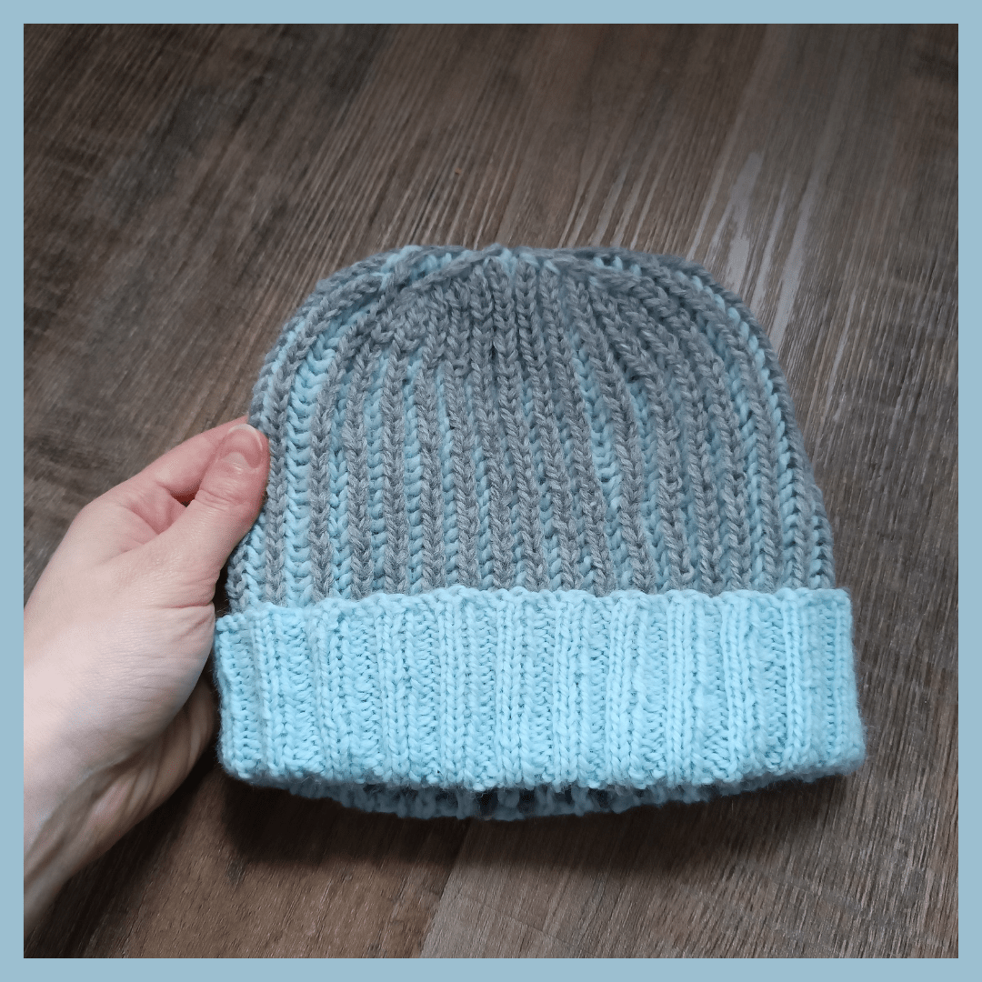 A light blue border surrounds a picture of a light blue and dark grey knitted hat. It's made in two coloured brioche. The main colour is dark grey and the turned up 2x2 ribbing is light blue. It's the same hat as in the first picture but inside out. The hat is held by a hand from the left side of the picture in front of a dark wooden floor.