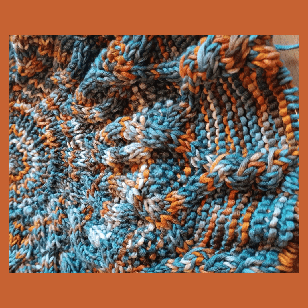 A close-up of a round knitted blanket. It's made with a hand-dyed yarn in blue, terracotta and white. The center of the blanket is on the left-hand side of the picture. There are cables spreading out towards the upper, right and under side. The needle with the live stitches can be made out on the right side.