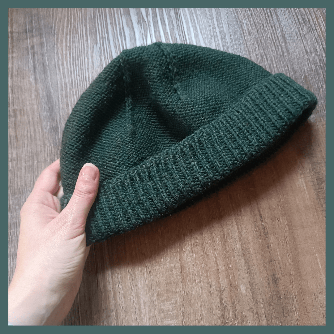 A dark green fisherman's toque is the center of the picture. A fair skinned hand holds it from the lower left side. The toque shows the purl side out with accented decreases at the top and a folded brim knitted with twisted stitches. The background is a dark wooden floor and there's a green frame around the picture.