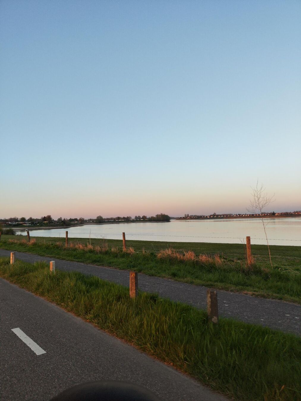 Dieses Bild zeigt eine friedliche Landschaft bei Sonnenuntergang.

- Im Vordergrund befindet sich ein asphaltierter Weg, der sich in die Ferne erstreckt. Der Weg ist mit weißen Markierungen versehen.
- Links und rechts des Weges befindet sich Gras, das von kleinen Pfosten begrenzt wird, die in regelmäßigen Abständen stehen.
- Hinter dem Weg erstreckt sich ein großes Gewässer, das ruhig daliegt und das Licht des Sonnenuntergangs reflektiert.
- Am Horizont sind Bäume und einige Gebäude zu sehen, die sich gegen den klaren Himmel abzeichnen.
- Der Himmel ist in sanften Farbtönen gehalten, die von Hellblau zu einem zarten Rosa übergehen, was auf den Sonnenuntergang hinweist.