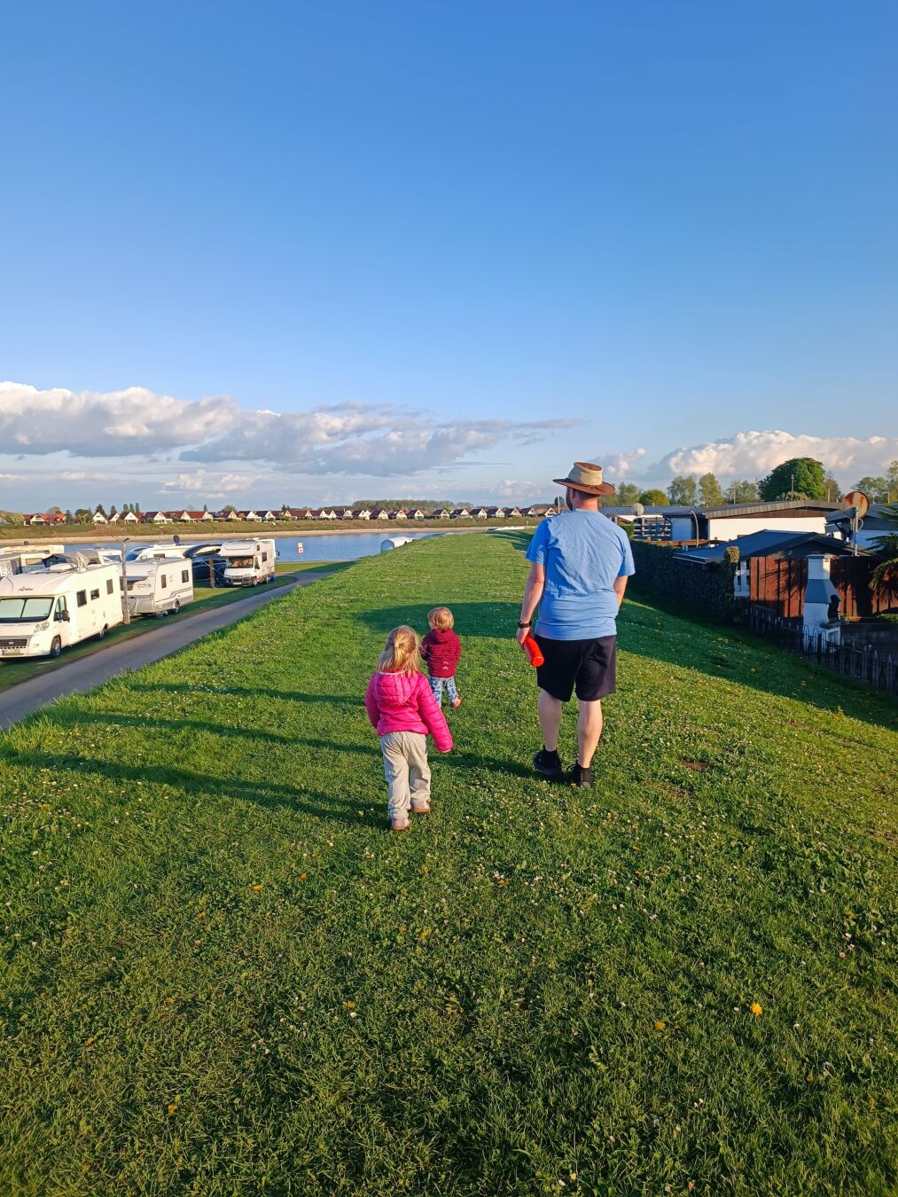 Ein sonniger Tag auf einem grünen, leicht abfallenden Hügel, hier ein Damm. Im Vordergrund gehen drei Personen mit dem Rücken zur Kamera den Damm entlang. Eine erwachsene Person in einem hellblauen T-Shirt, dunklen Shorts und einem hellen Hut geht neben zwei Kindern. Das Kind direkt hinter der erwachsenen Person trägt eine leuchtend pinke Jacke und helle Hosen. Das kleinste Kind trägt eine rote Jacke und helle Jeans. Links im Bild stehen mehrere weiße Wohnmobile auf einer befestigten Fläche. Dahinter erstreckt sich ein Gewässer unter einem hellblauen Himmel mit einigen weißen Wolken. Am gegenüberliegenden Ufer sind mehrere kleine Ferienhäuser zu sehen. Rechts im Bild befinden sich Mobilheime und grüne Vegetation.