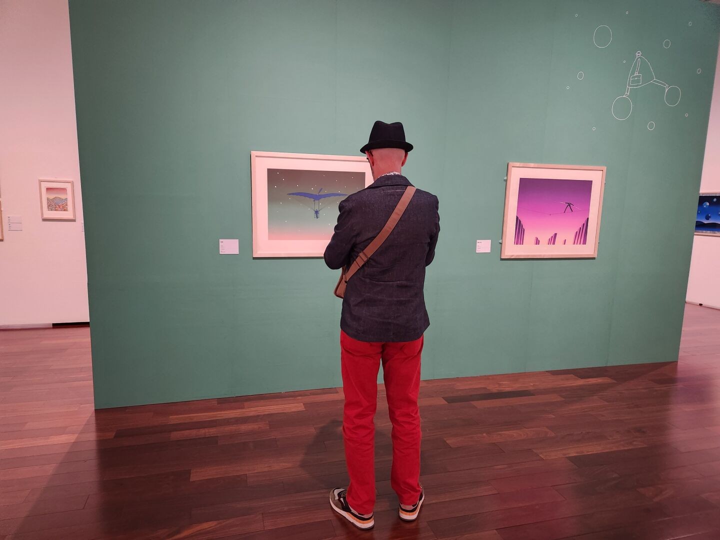 A man wearing a dark blue denim hat, dark blue denim blazer, red denim pants, and sneakers is standing in an art gallery, looking at framed artworks by Folon displayed on a green wall. The paintings feature surreal scenes with minimalist, colorful backgrounds. The gallery has wooden flooring and clean, modern decor.