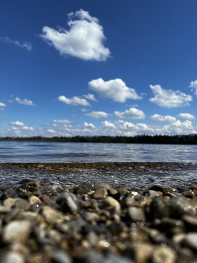 a photo in portrait mode, in the foreground some wet gravel with a like behind, in the distance a shoreline with trees and above that blue skies with some random white clouds. All very lively and with high contrast.