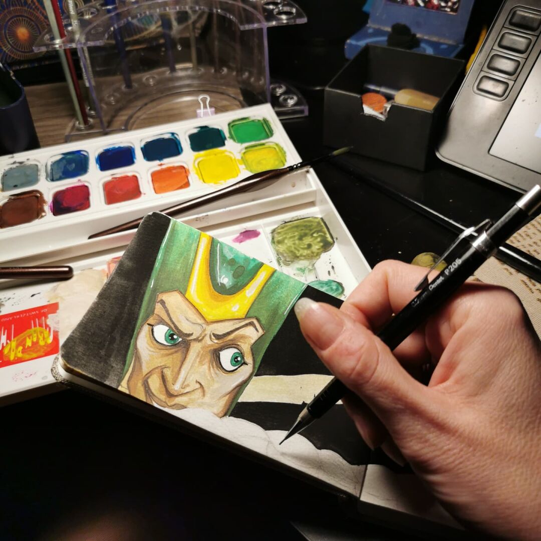 Wider foto of: Foto of a sketchbook page. A male face with brown skun, green eyes and an Egyptian styke hat is to be seen. Also my fingers with an ink pen and my colors.