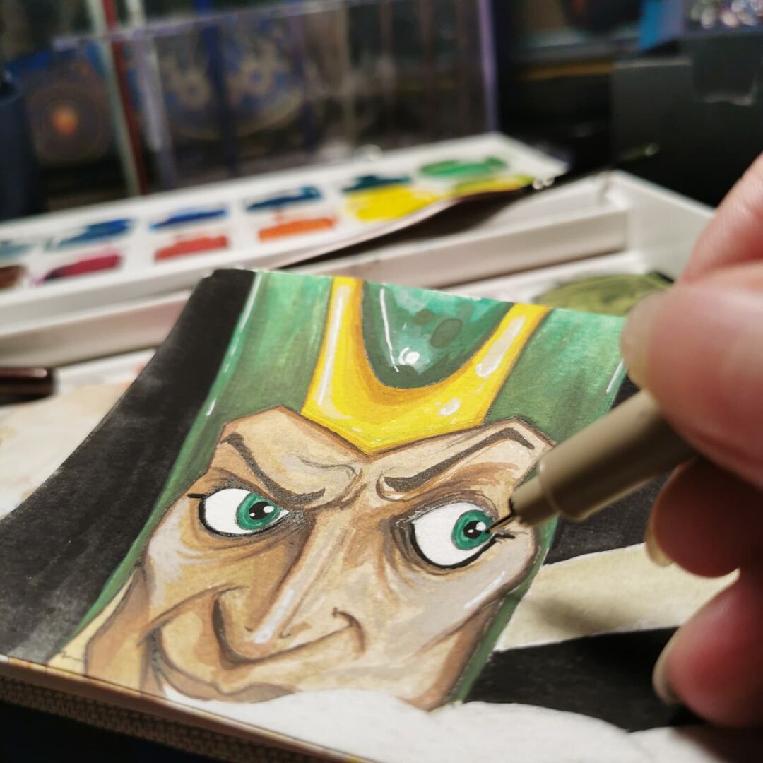 Foto of a sketchbook page. A male face with brown skun, green eyes and an Egyptian styke hat is to be seen. Also my fingers with an ink pen and my colors.