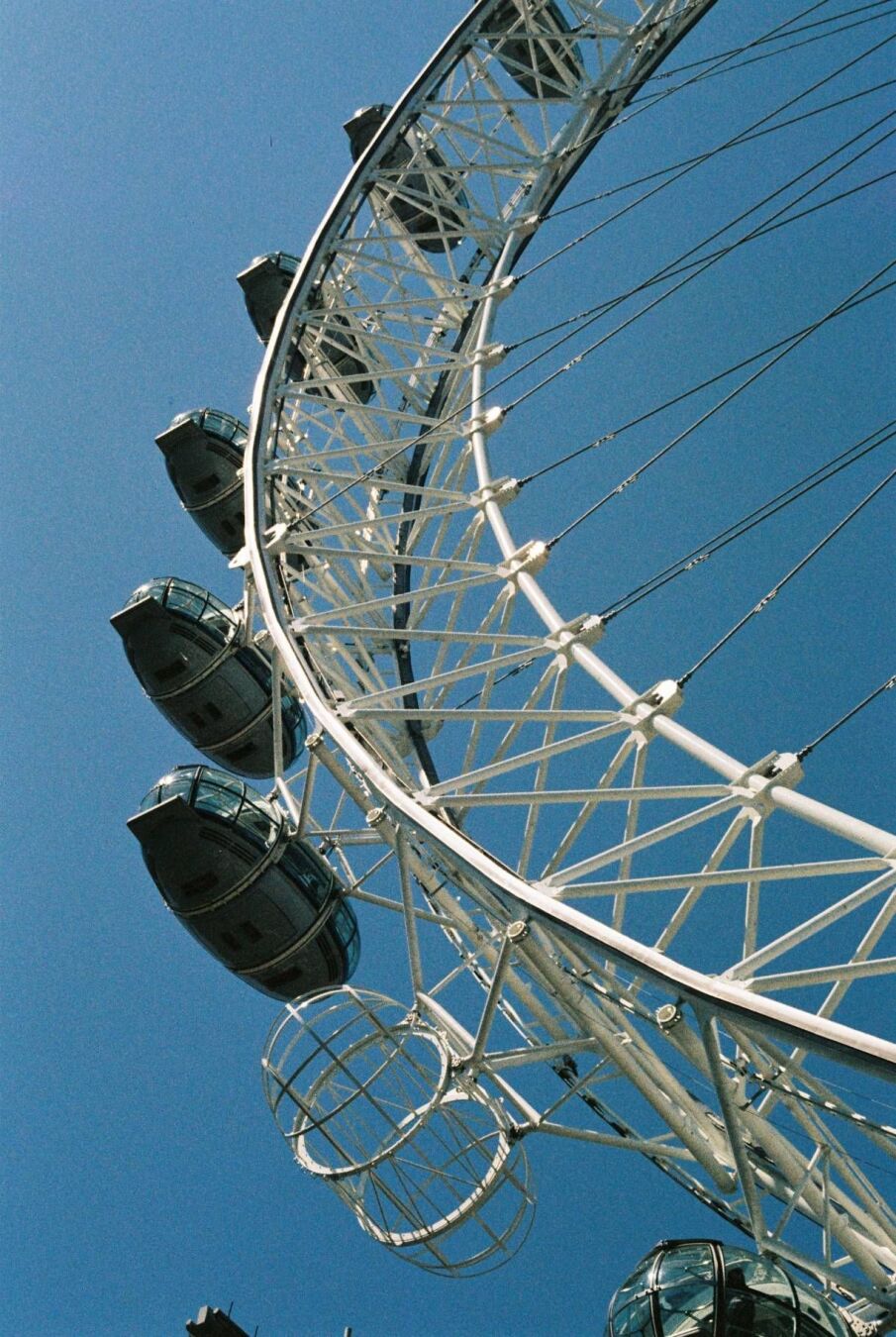 The London Eye seen from below at an angle, its capsules mounted along the white steel rim, thin spokes and cables radiating out to the hub. The sky is a clear, deep blue with no clouds, the light hard and contrasty, giving the structure crisp, sharp edges.